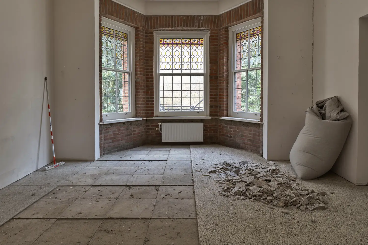 Voorkamer van een jaren dertig woning met glas-in-loodramen, waar de helft van de oude vloertegels al is verwijderd en de kale dekvloer zichtbaar is